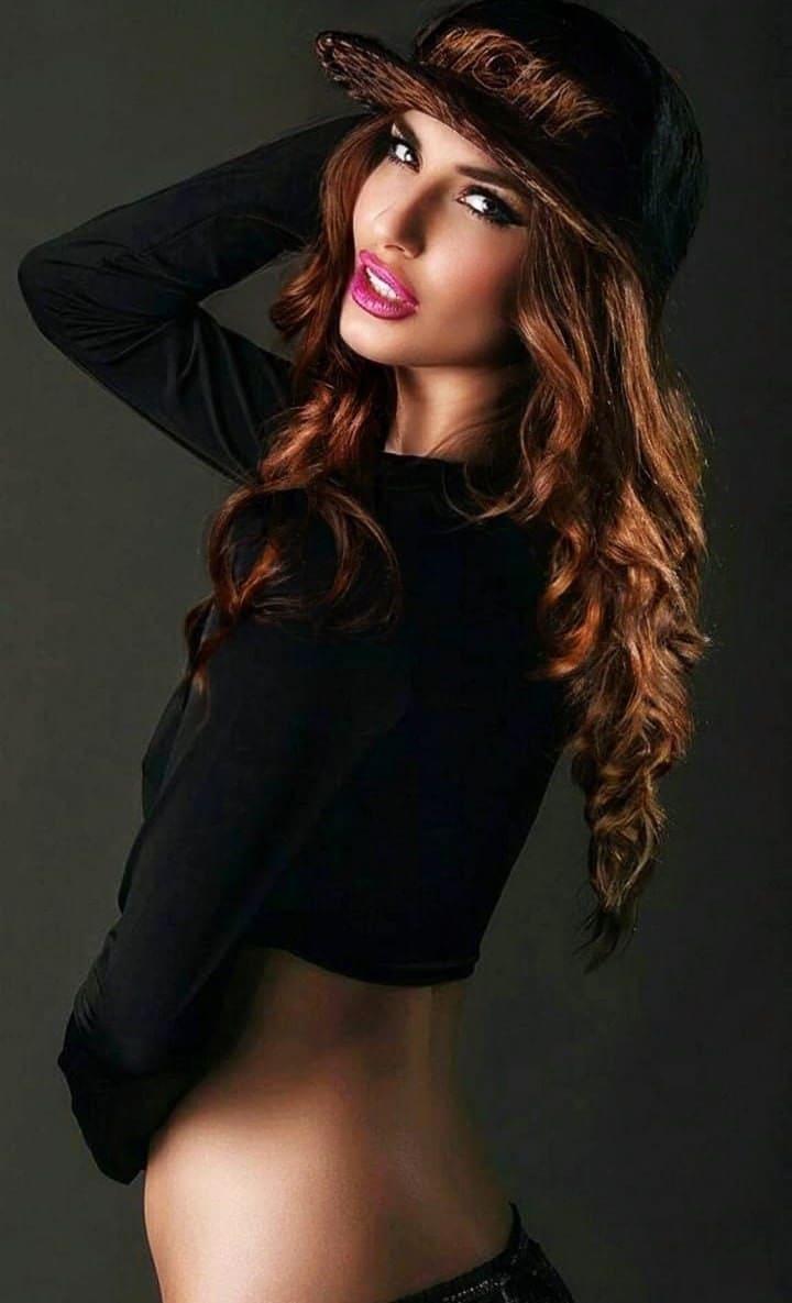 A woman in a black cap and long-sleeved crop top poses with one arm raised behind her head against a dark gray studio background.