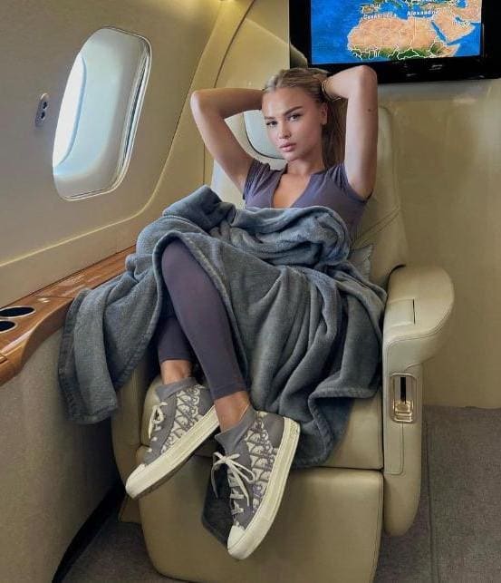 Blonde woman relaxes in a private jet seat wearing a grey top, leggings, and sneakers, with a blanket over her legs.