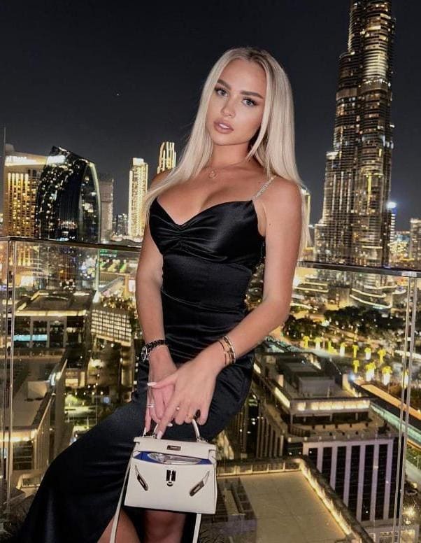 Woman in a black satin slip dress sits on a rooftop ledge at night, holding a white handbag against a glowing skyline.