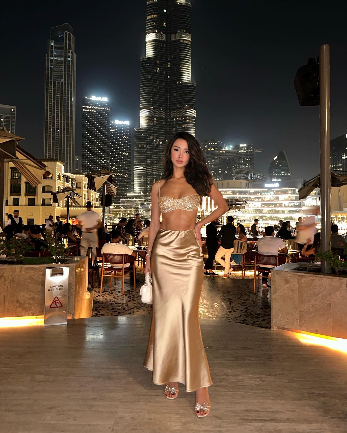 A woman in a gold sequined top and satin skirt stands on a terrace at night, framed by lit skyscrapers in the background.
