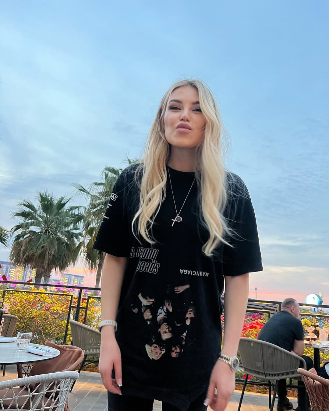 A young blonde woman stands on an outdoor patio wearing an oversized black graphic t-shirt, with palm trees and a railing behind her.