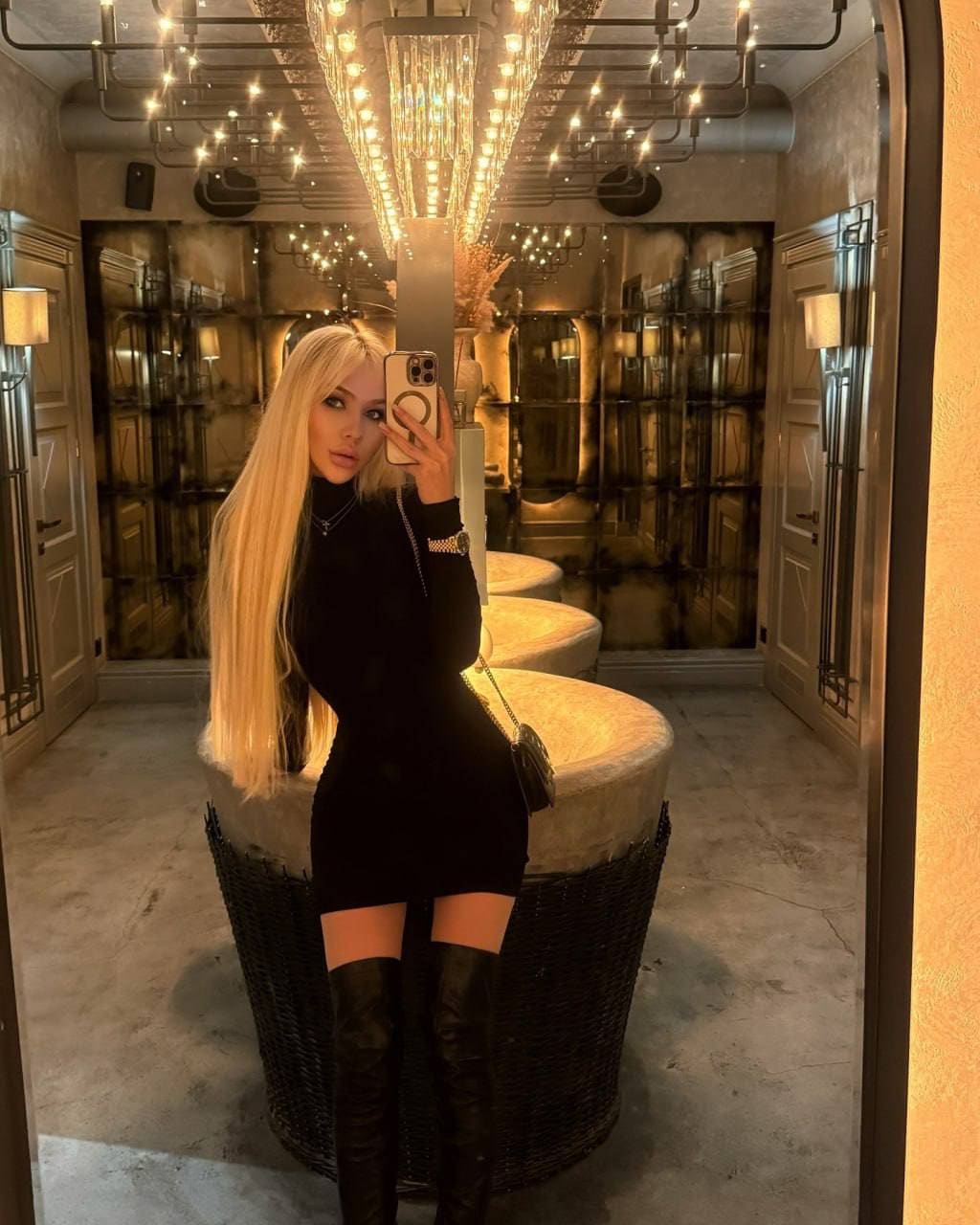 A woman in a black mini dress and thigh-high boots takes a mirror selfie near illuminated sinks under a chandelier-lit ceiling.