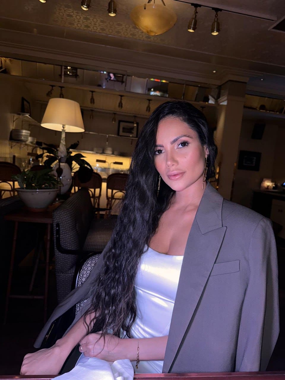 Sophie sits at a restaurant table in a white satin top and open grey blazer, with a lamp and chairs in the dimly lit background.