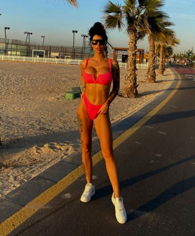 A woman in a pink bikini and white sneakers stands on a paved beach path, wearing sunglasses with palm trees behind her.