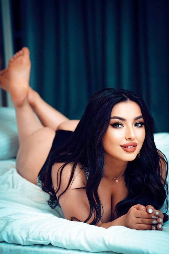 Chantal lies on her stomach on a light bed, wearing a black strapless top and necklace, with legs raised against a dark curtain.