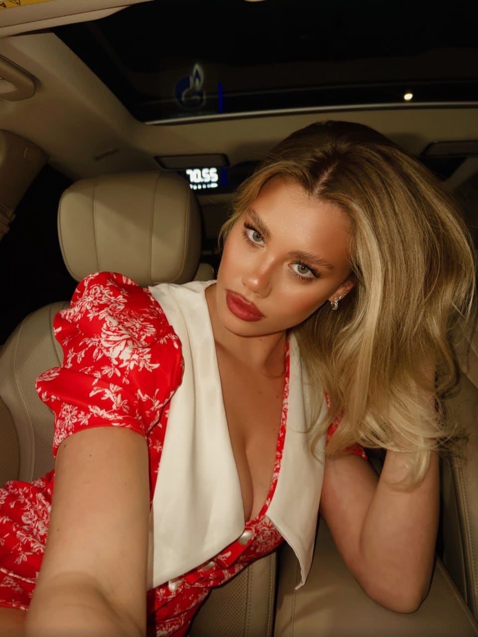 A blonde woman in a red floral dress with a white collar sits in the back seat of a car, looking at the camera.