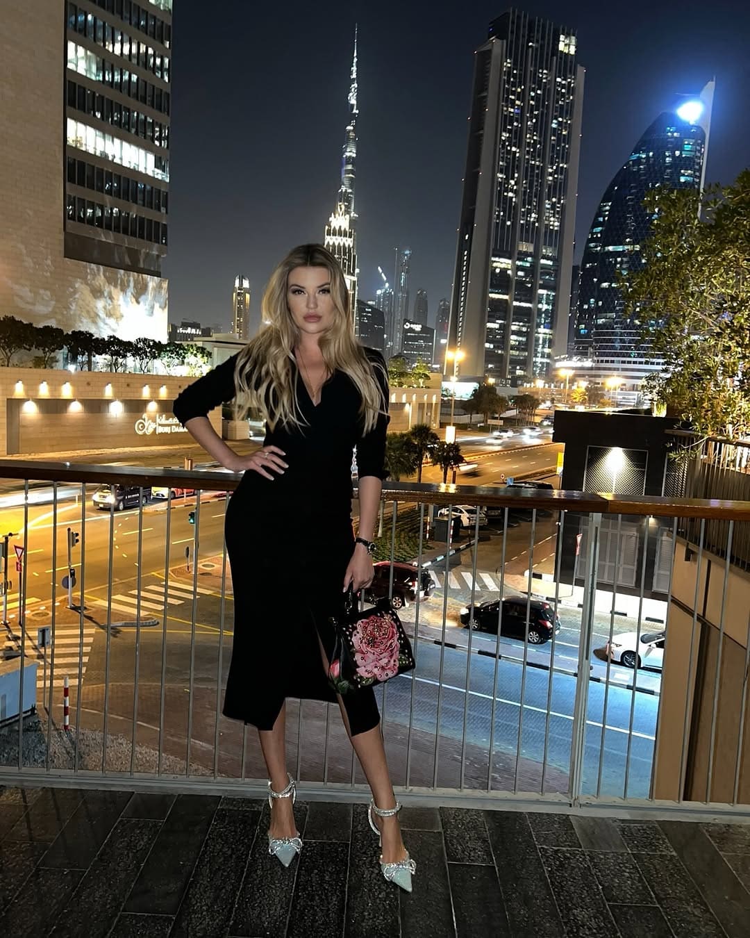 A woman in a black knee-length dress with a slit stands on a balcony at night, holding a floral handbag against a city skyline backdrop.