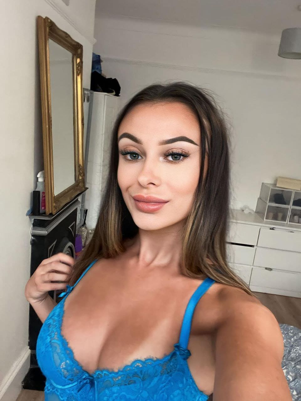 Tia stands in a bedroom in a blue lace camisole, taking a selfie before a gold-framed mirror and white dresser.