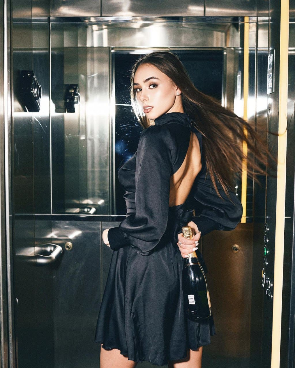 A woman with dark hair looks over her shoulder in a metallic elevator, wearing a backless black dress and holding a champagne bottle behind her back.