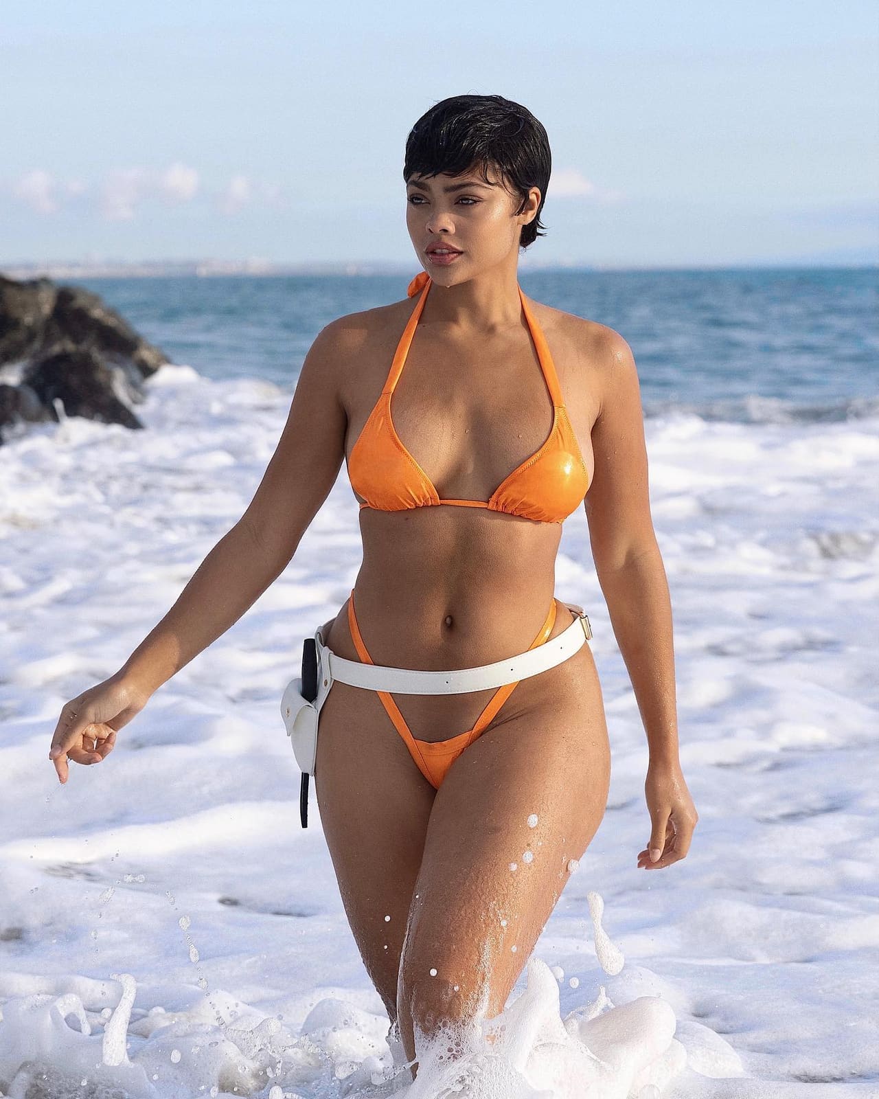 A woman in an orange halter bikini walks through foamy surf on a rocky shoreline under a bright sky.