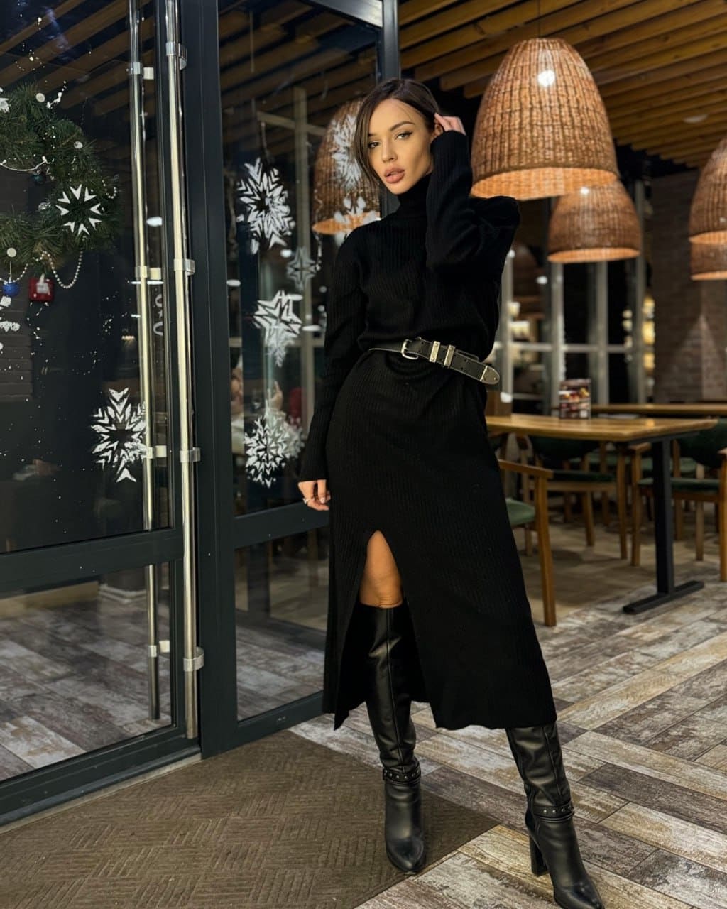 A woman in a black turtleneck dress with a slit and knee-high boots stands before a storefront with snowflake decals.