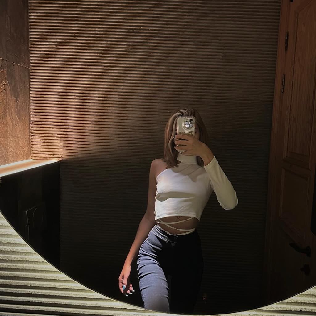 A woman takes a mirror selfie in a white one-shoulder crop top and dark jeans, standing in front of a ribbed wooden wall.