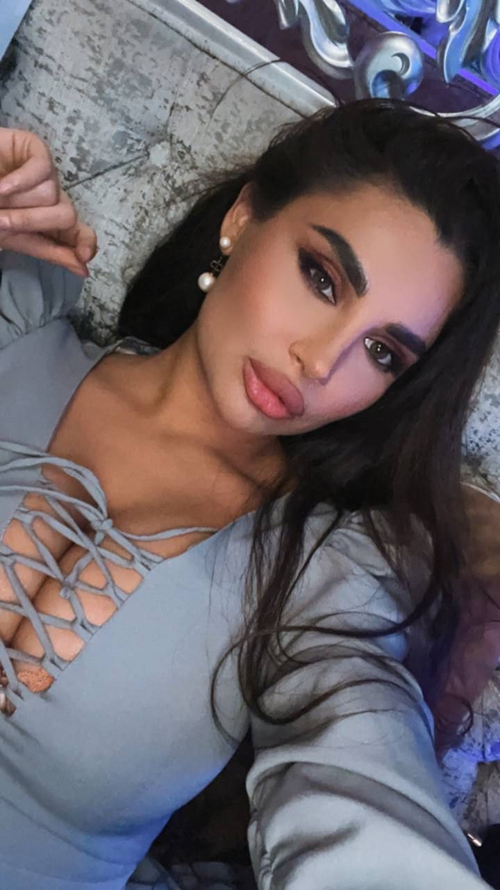 A woman lies on a textured surface wearing a grey lace-up top and pearl drop earrings, with the front open revealing part of her chest.
