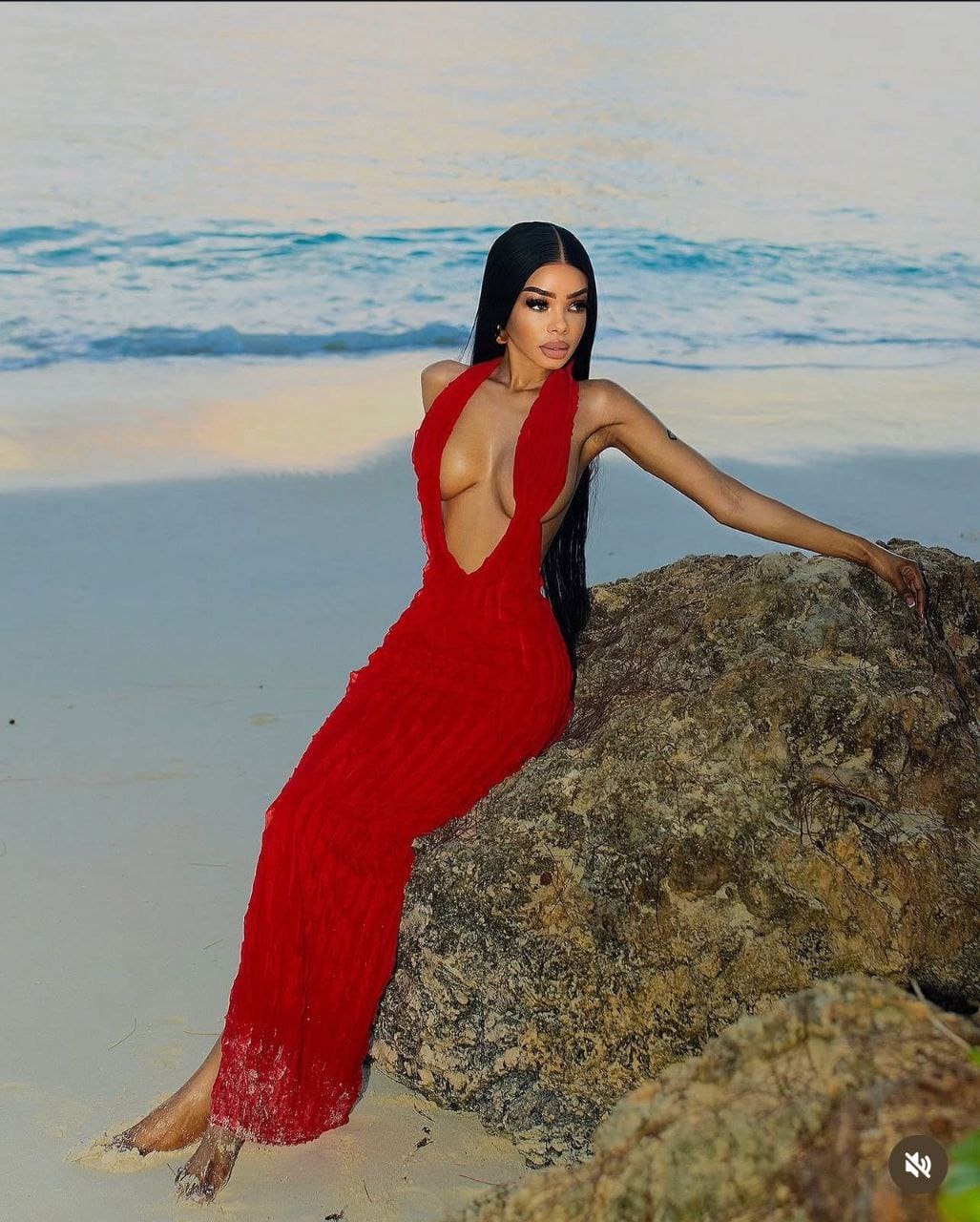 A woman sits on a beach rock wearing a long red dress with a deep neckline, the ocean and waves stretching out behind her on the sandy shore.