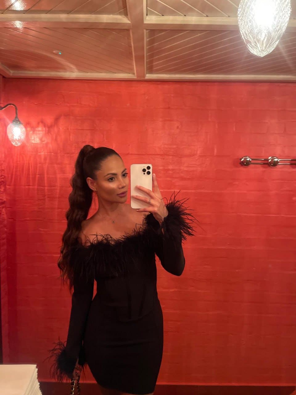 A mirror selfie shows a woman in a black off-the-shoulder mini dress with feathered trim, holding a white phone in a room with red brick walls.