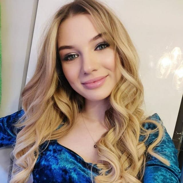 Close-up selfie of a woman in a blue velvet top and necklace against a plain light-colored wall.