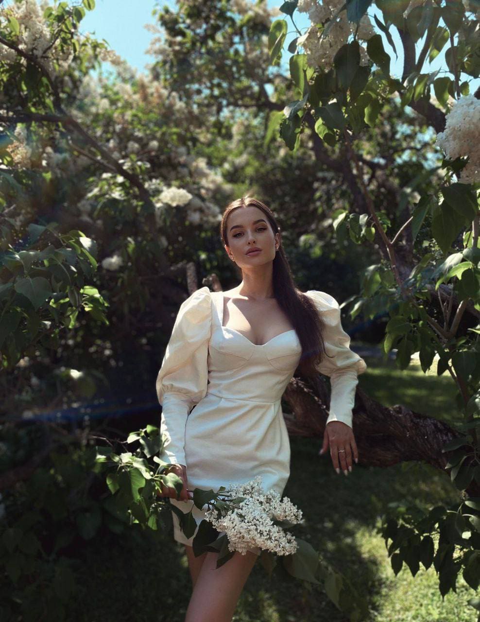 A woman in a cream dress with puffed shoulders stands among flowering trees, holding a branch of white blossoms.