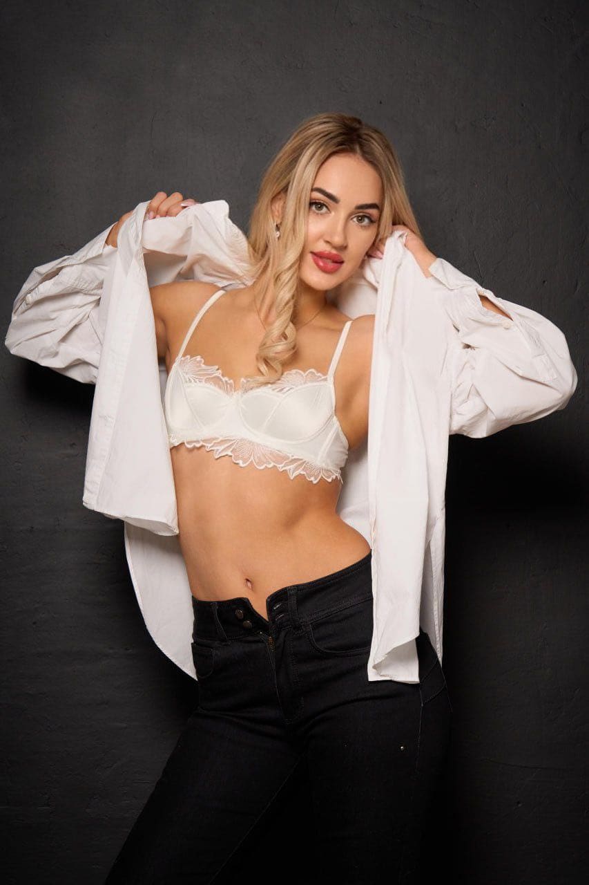 Trinity stands against a gray wall, opening a white shirt to reveal a lace bra and bare midriff while wearing black jeans.