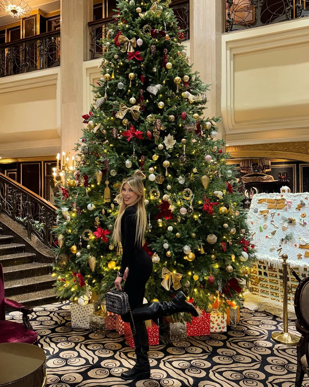 A blonde woman poses with one leg raised in a grand lobby, wearing a black bodysuit and knee-high boots while holding a patterned handbag.