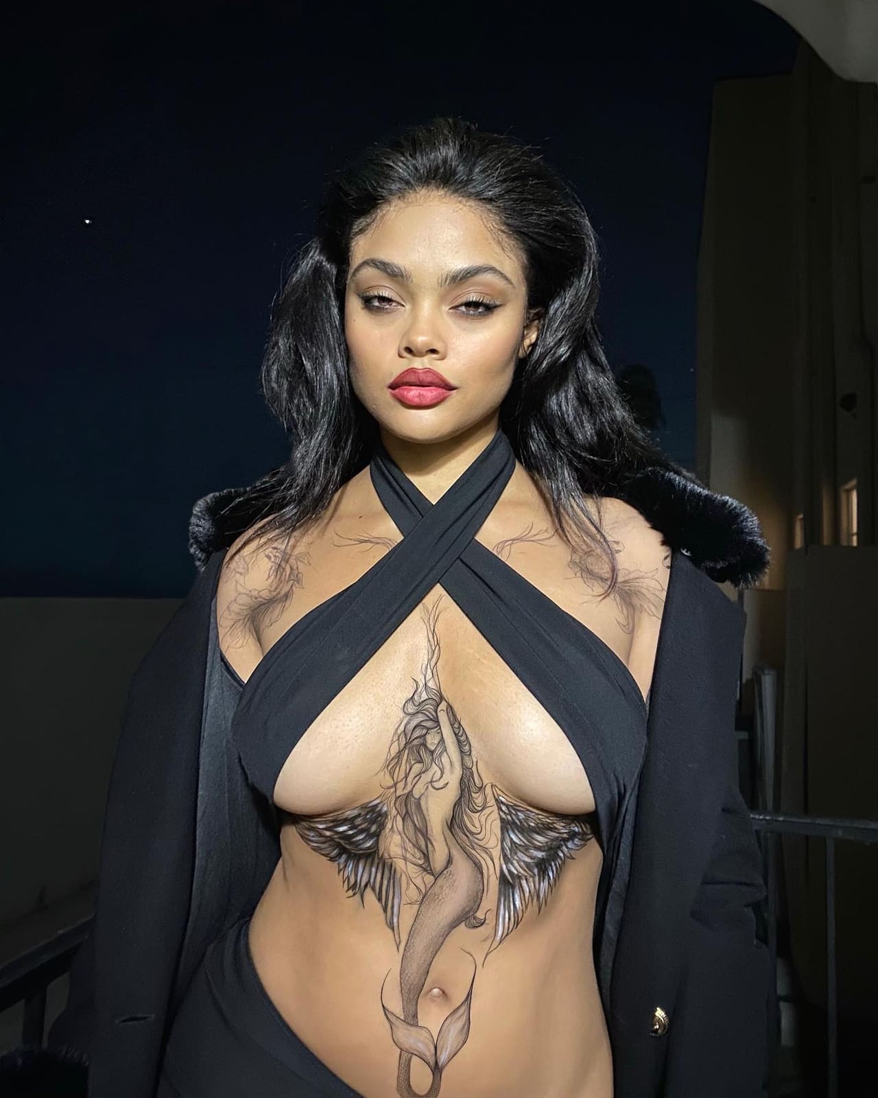 A woman in a black cross-halter top and fur-collared jacket stands at night, revealing chest and abdomen tattoos.