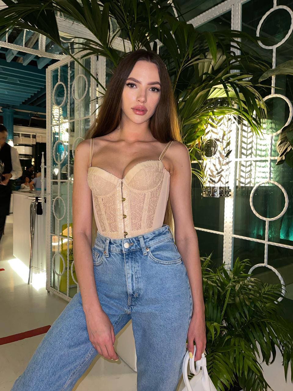 Polya stands indoors in a beige lace corset and light blue jeans, holding a white handbag near green plants and white lattice panels.