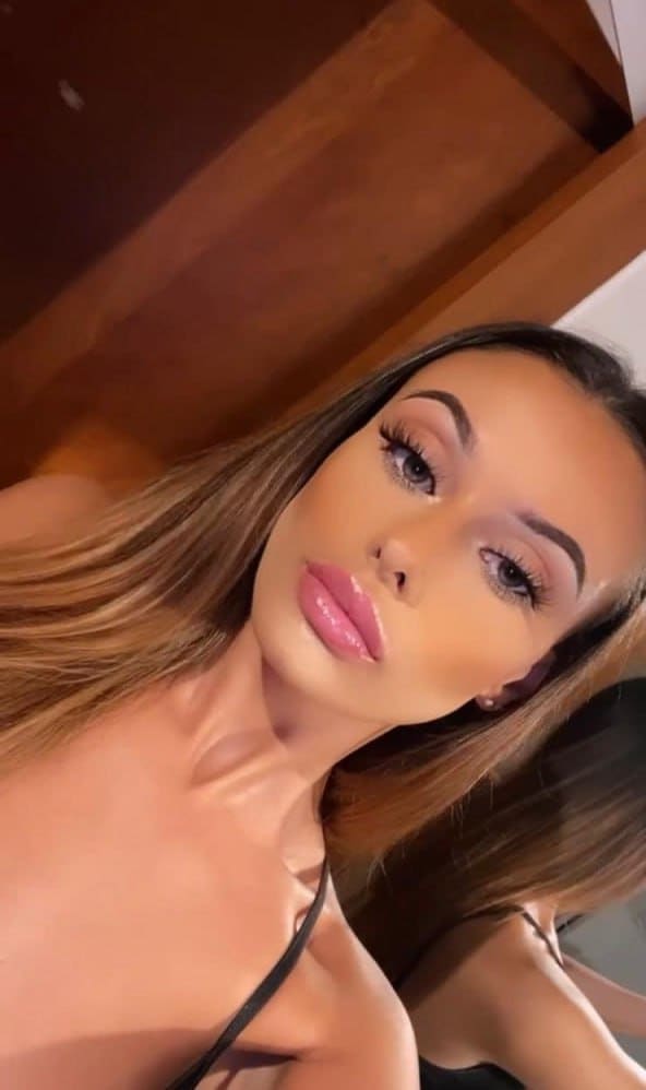 Close-up selfie of a woman with long brown hair and pink lips, wearing a black strap top against a wooden background.