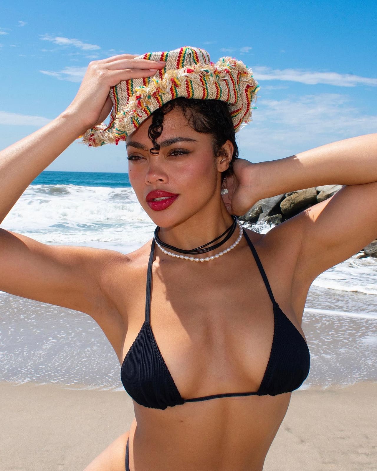 A woman in a black string bikini and woven hat stands on a beach with one arm raised, holding the hat with the other.