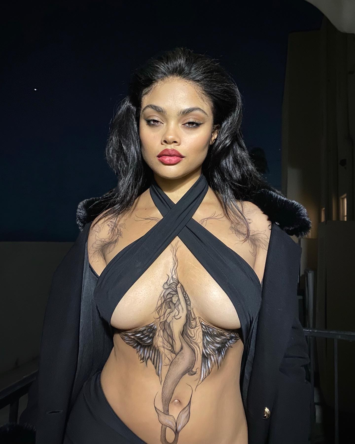 A woman in a black cross-halter top and fur-collared jacket stands at night, revealing chest and abdomen tattoos.
