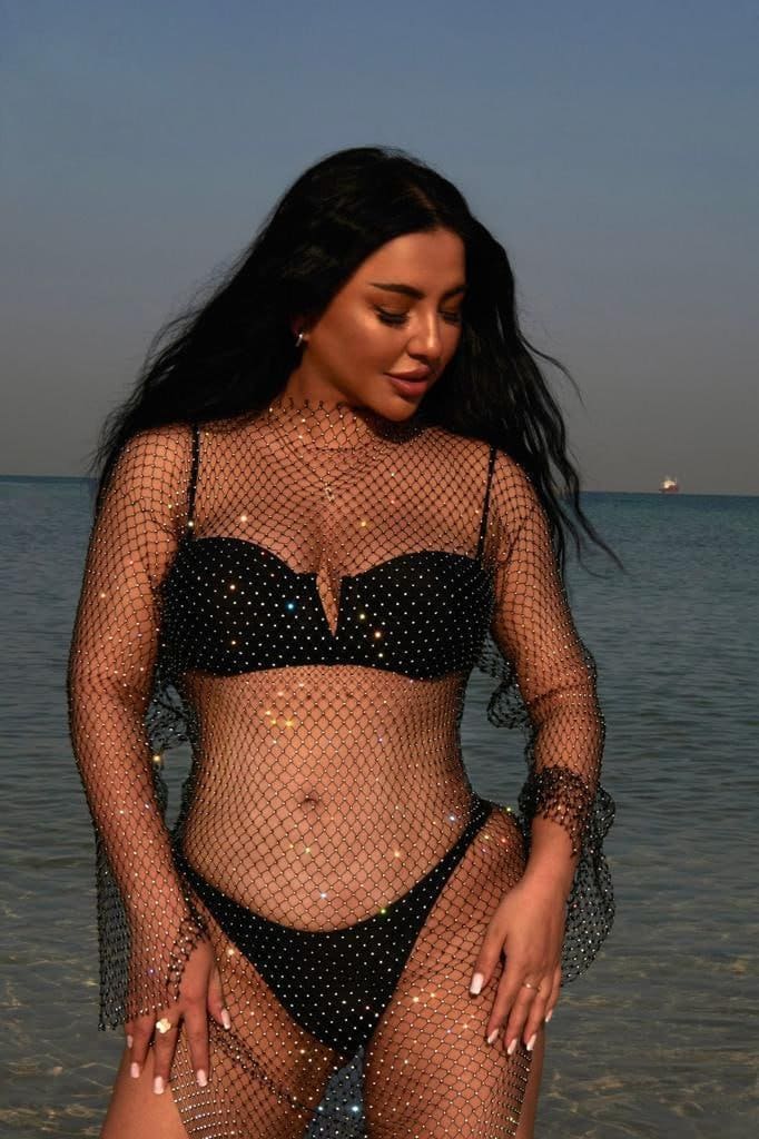 A woman at the water's edge wears a black studded bikini under sheer sparkly mesh netting, head tilted down with hands on thighs.