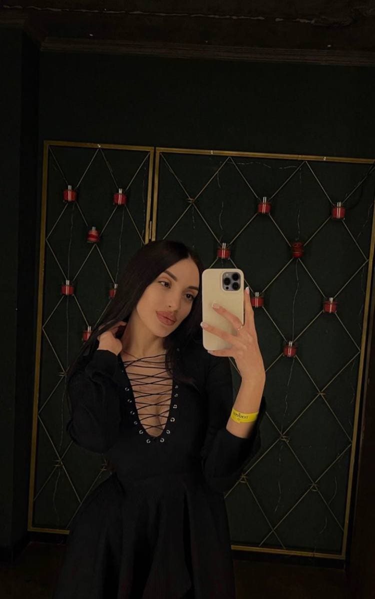 She takes a mirror selfie in a black long-sleeved lace-up dress, standing before a dark paneled wall illuminated by small red lights.