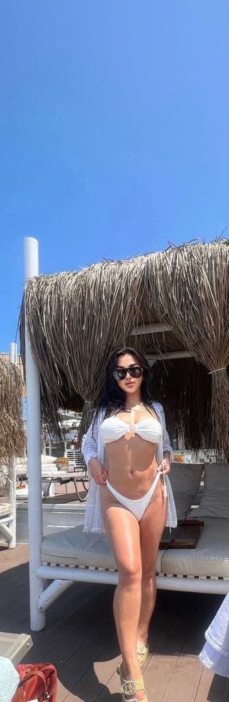 A woman in a white string bikini and open cover-up stands on a wooden deck before a thatched cabana under a clear blue sky.