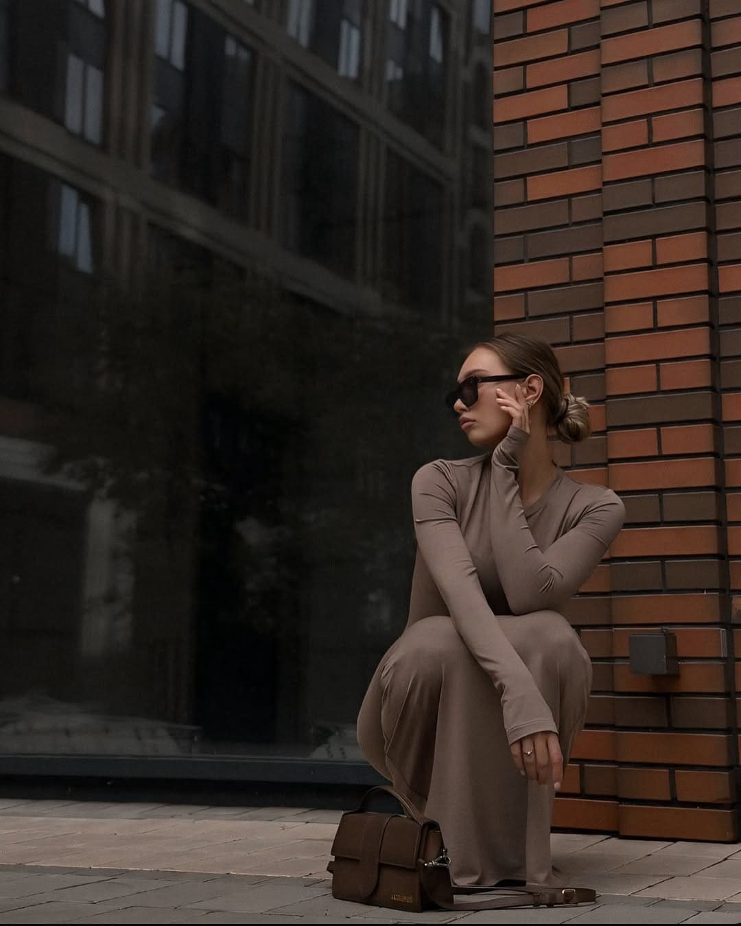 A woman in a taupe turtleneck dress and sunglasses crouches on a sidewalk beside a brick wall and glass window.