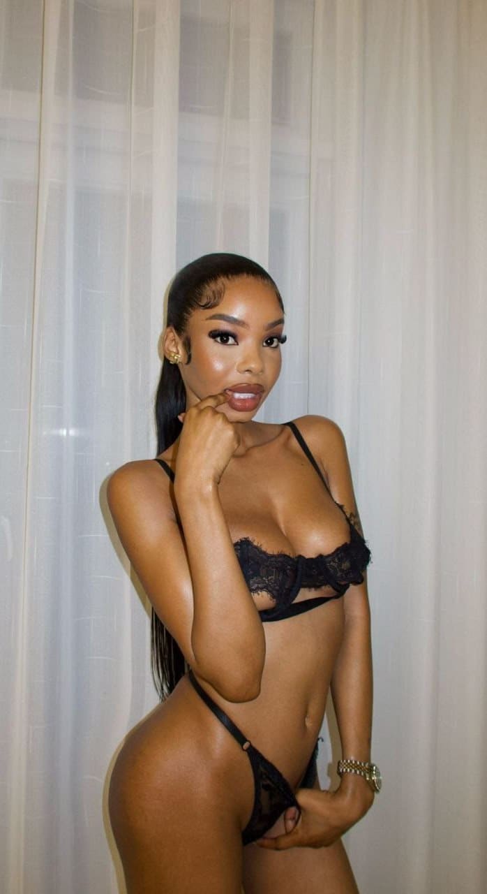 A woman in black lace lingerie stands before sheer curtains, one hand raised to her chin and the other near her hip, with long dark hair pulled back.