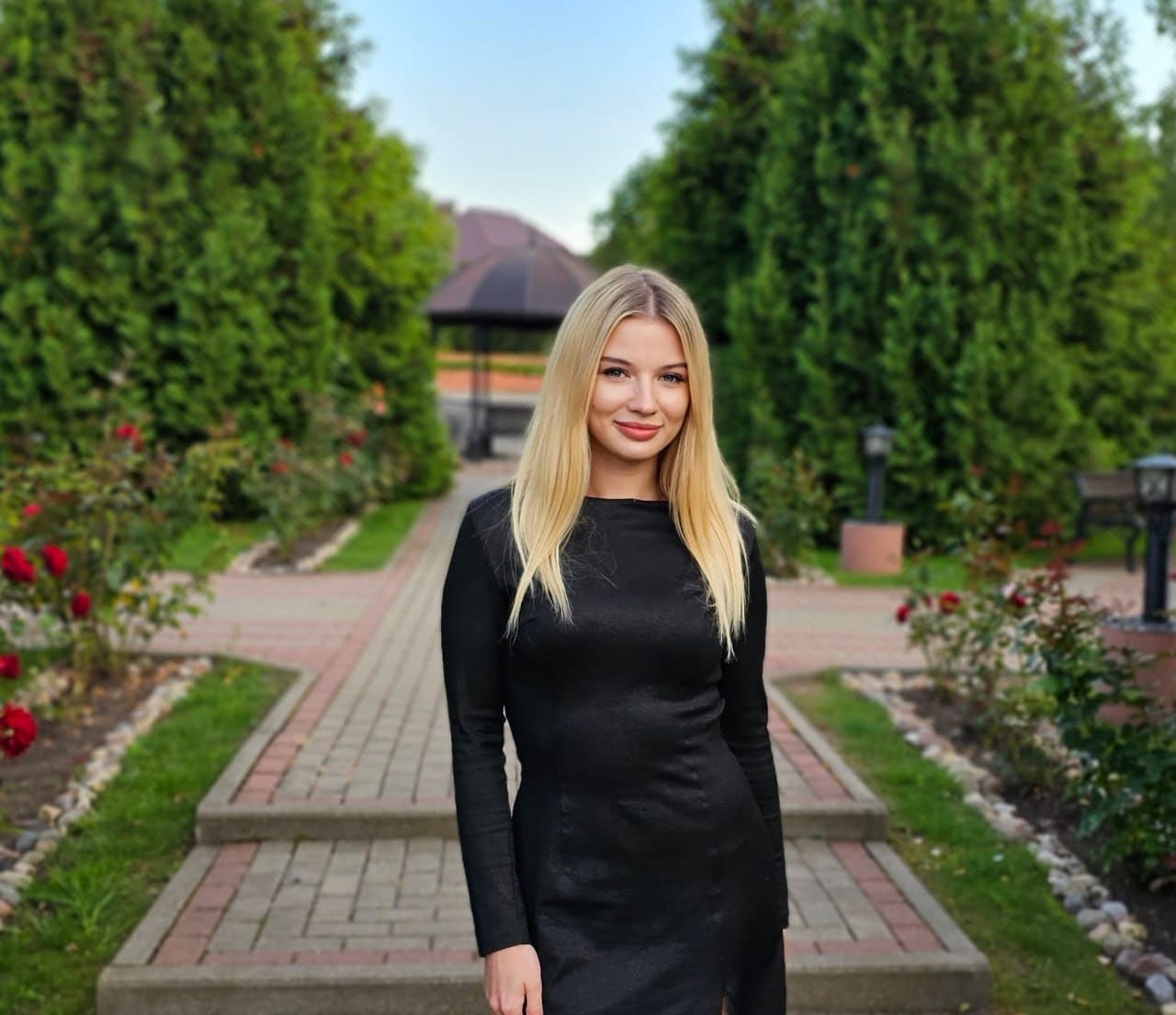 Daria stands in a black long-sleeved dress on a brick path with hedges, roses, and a gazebo behind her.