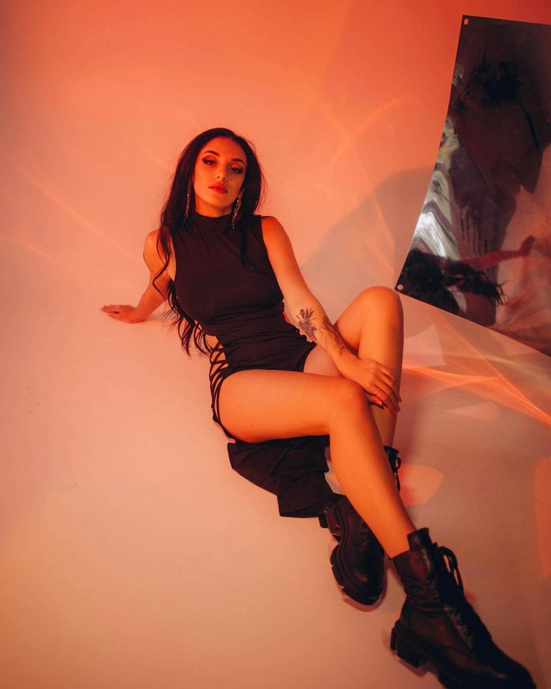 A woman with long dark hair sits on a white floor in a black side-laced mini dress and lace-up boots, with a mirror to her right.