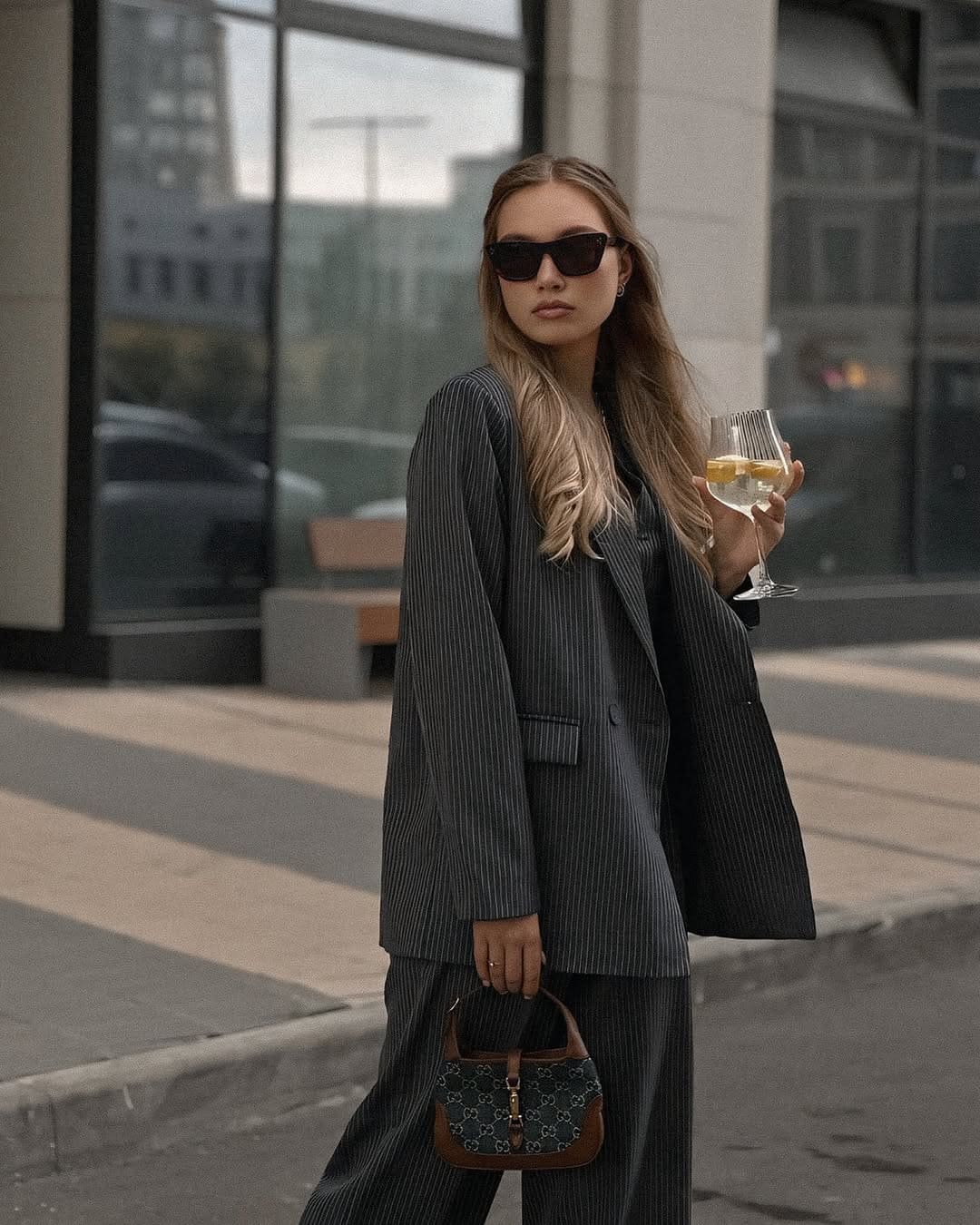 A woman in a pinstriped suit and sunglasses stands on a sidewalk holding wine and a handbag near a glass building.