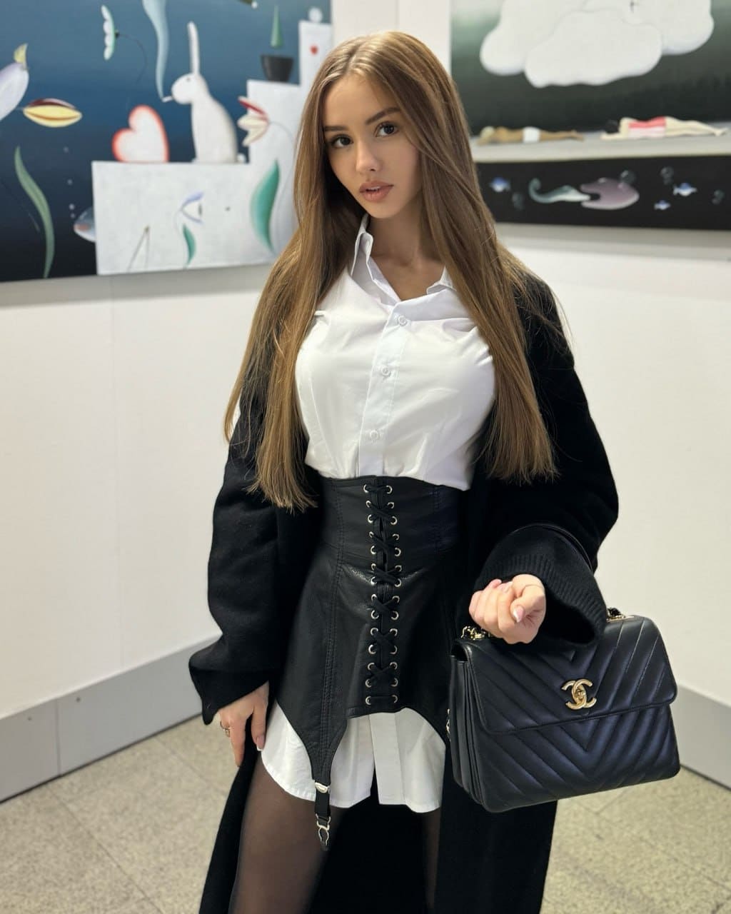 Sophia stands indoors in a white shirt, black corset, coat, and tights, holding a handbag before colorful artwork.