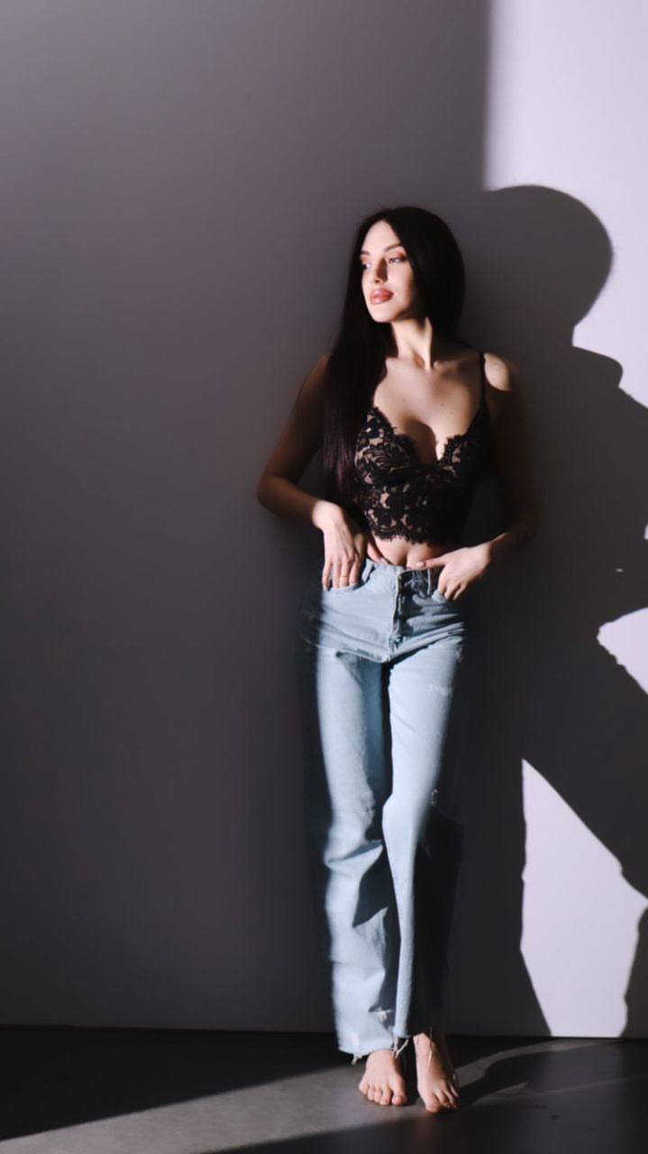 Lika stands barefoot against a plain wall in a black lace camisole and light blue jeans, hands on hips, casting a strong shadow.