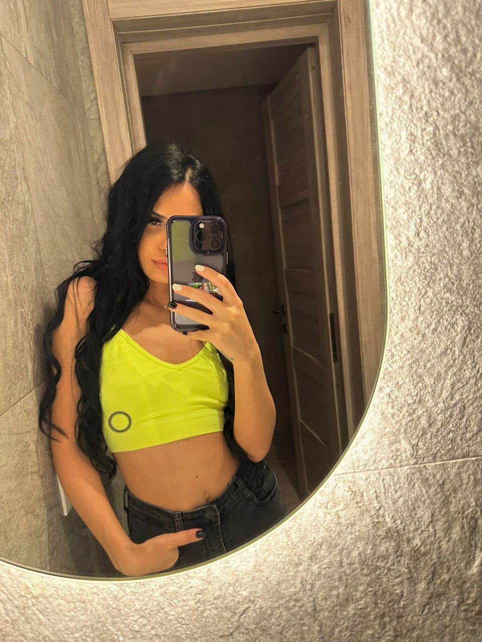 A woman in a lime-green cropped tank top and dark jeans takes a mirror selfie in a bathroom, holding a smartphone in front of her face.