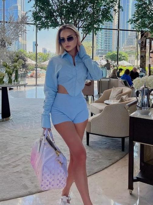 Blonde woman in a light blue cut-out outfit and sunglasses stands in a sunlit lounge holding a patterned handbag.