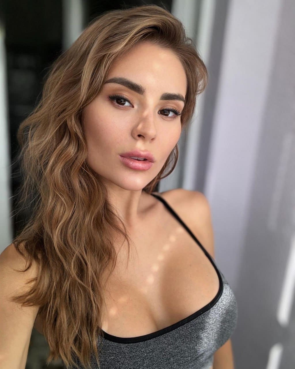 Fiona with long wavy light-brown hair wears a silver glittery top with thin black straps, posing indoors against a soft gray background.