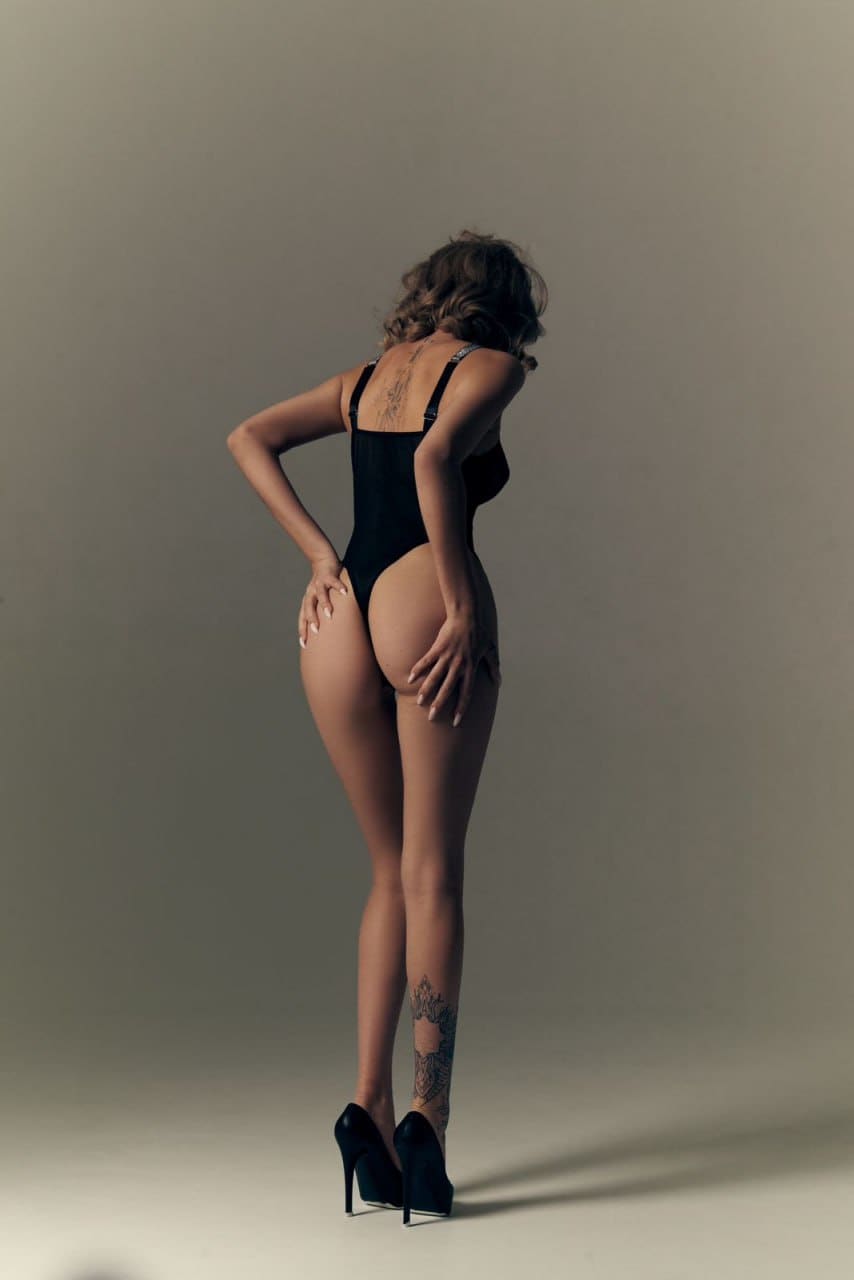 A woman with curly hair stands in profile against a gray backdrop, wearing a black bodysuit and heels, with hands resting on her hips and thighs.