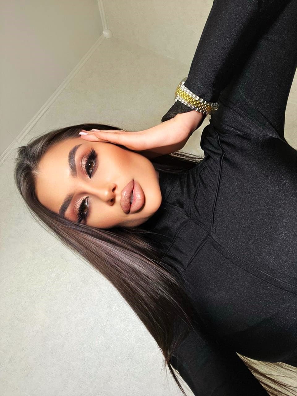 A woman lies on a light floor in a black long-sleeved top, wearing a bracelet, with one hand resting near her head.
