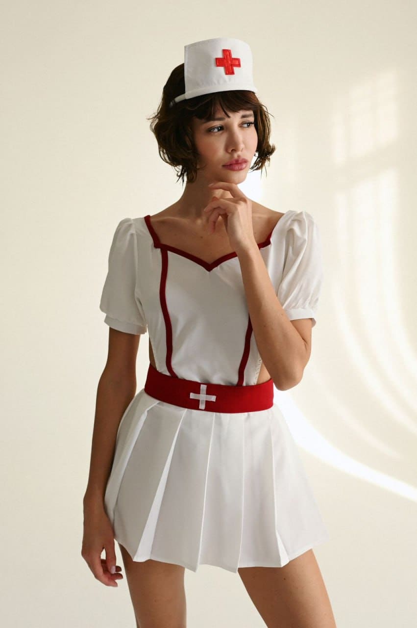 Viki stands in a studio wearing a white nurse costume with red trim and cap, posing with one hand near her chin.