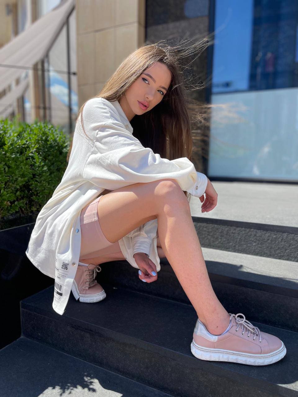 A woman crouches on stone steps in an oversized white shirt, pink skirt, and platform sneakers, with greenery and a building behind.
