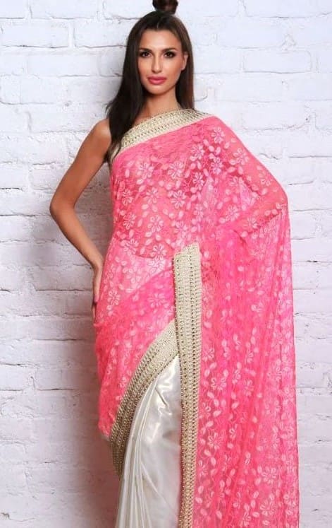 Standing against a white brick wall, she wears a pink floral sari with gold trim over a white blouse, her hand resting on her hip.