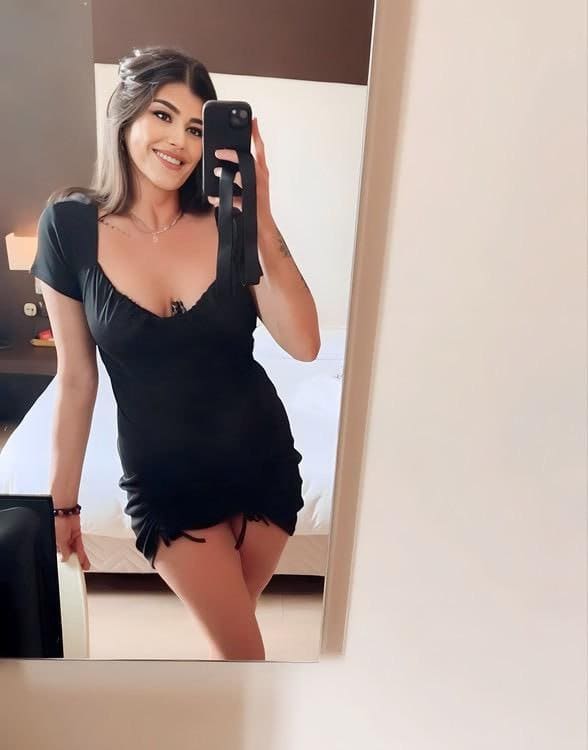 Milana takes a mirror selfie in a bedroom, wearing a short black dress with a deep neckline and holding a phone.