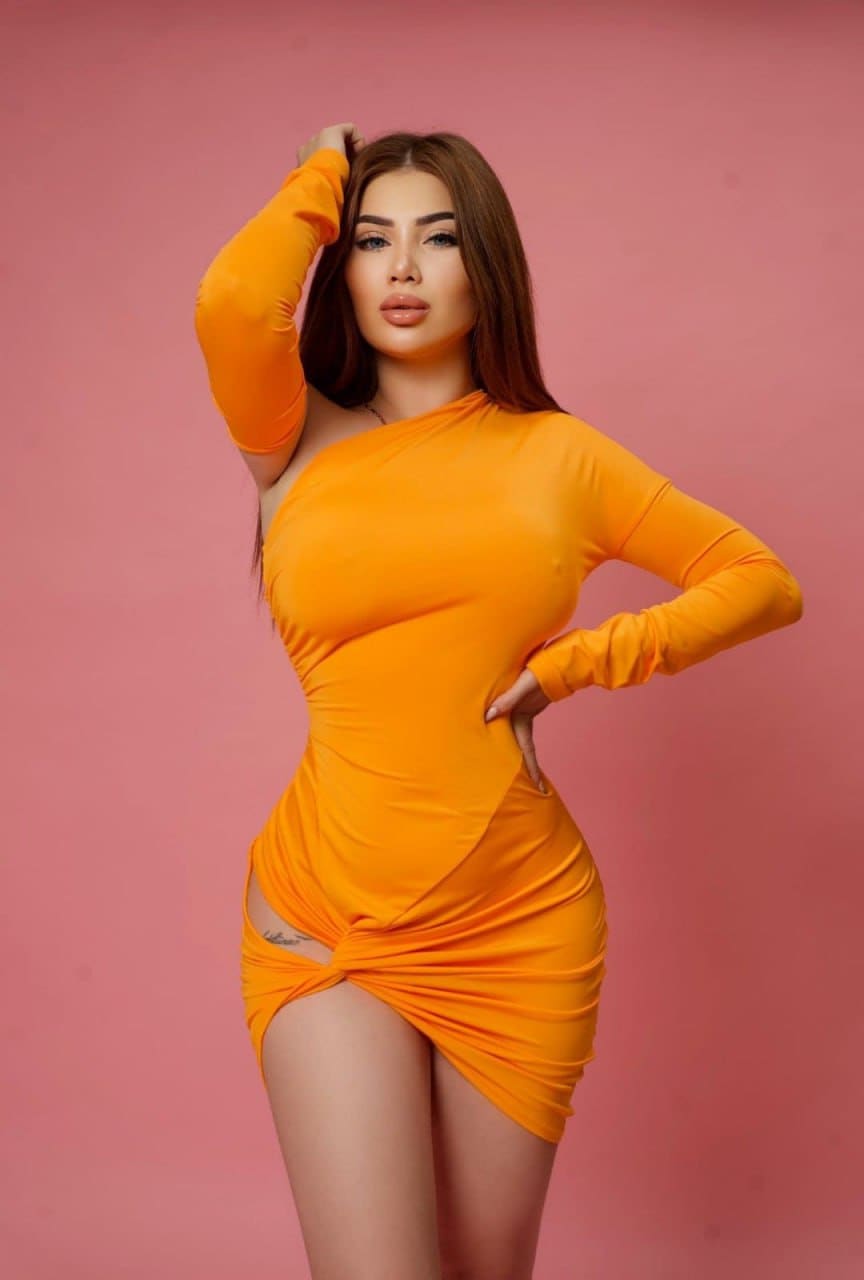 Lena stands against a pink backdrop in a tight orange mini dress with a high side slit, one hand on her hip and the other raised to her head.