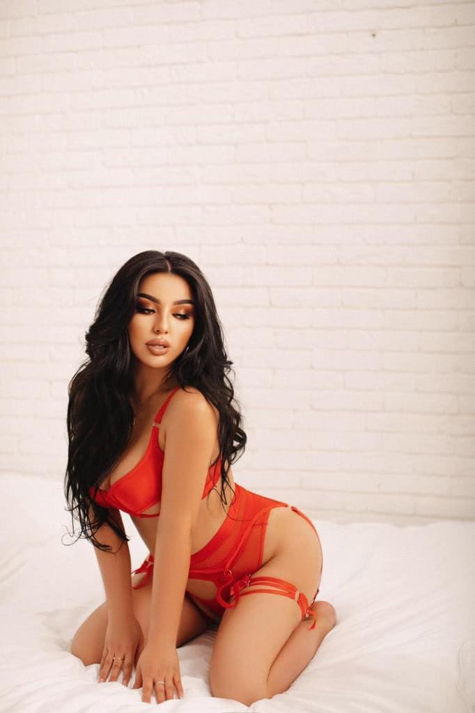 A woman kneels on a white bed before a white brick wall, wearing red strappy lingerie with a garter belt and hands on the sheets.