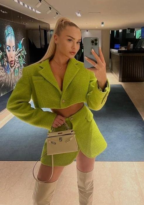Mirror selfie of a blonde woman in a green tweed jacket and mini skirt, beige boots, and handbag, posing with a phone.